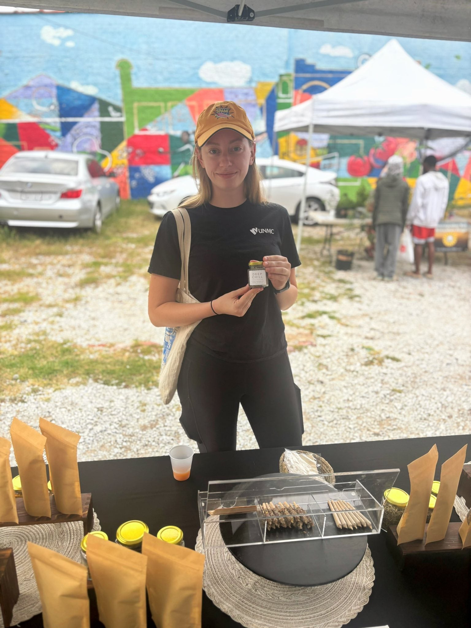 Woman in black shirt and yellow cap holding a Nava Living Co product at an outdoor event with colorful tents and cars in the background.