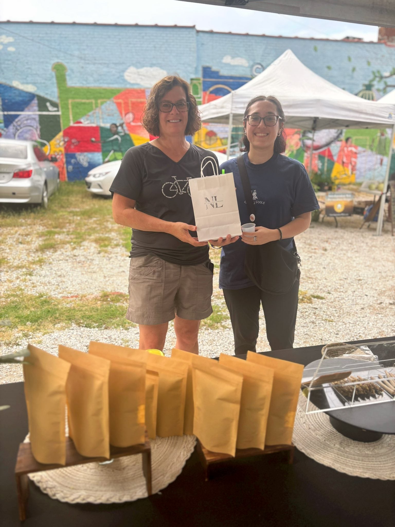 Two women standing outdoors with a table of Nava Living Co. herbal tea packages and a colorful mural in the background.