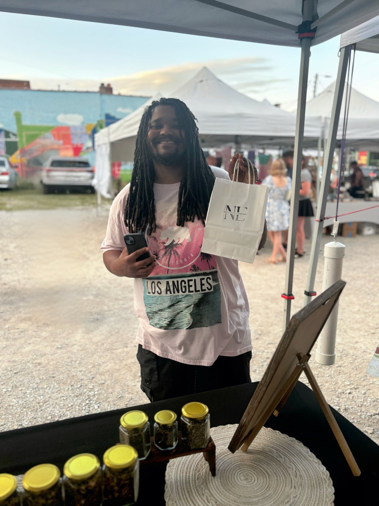 Kenny Lacey holding a phone and a Nava Living Co. bag, standing under a tent with loose leaf tea jars on a table in the foreground.