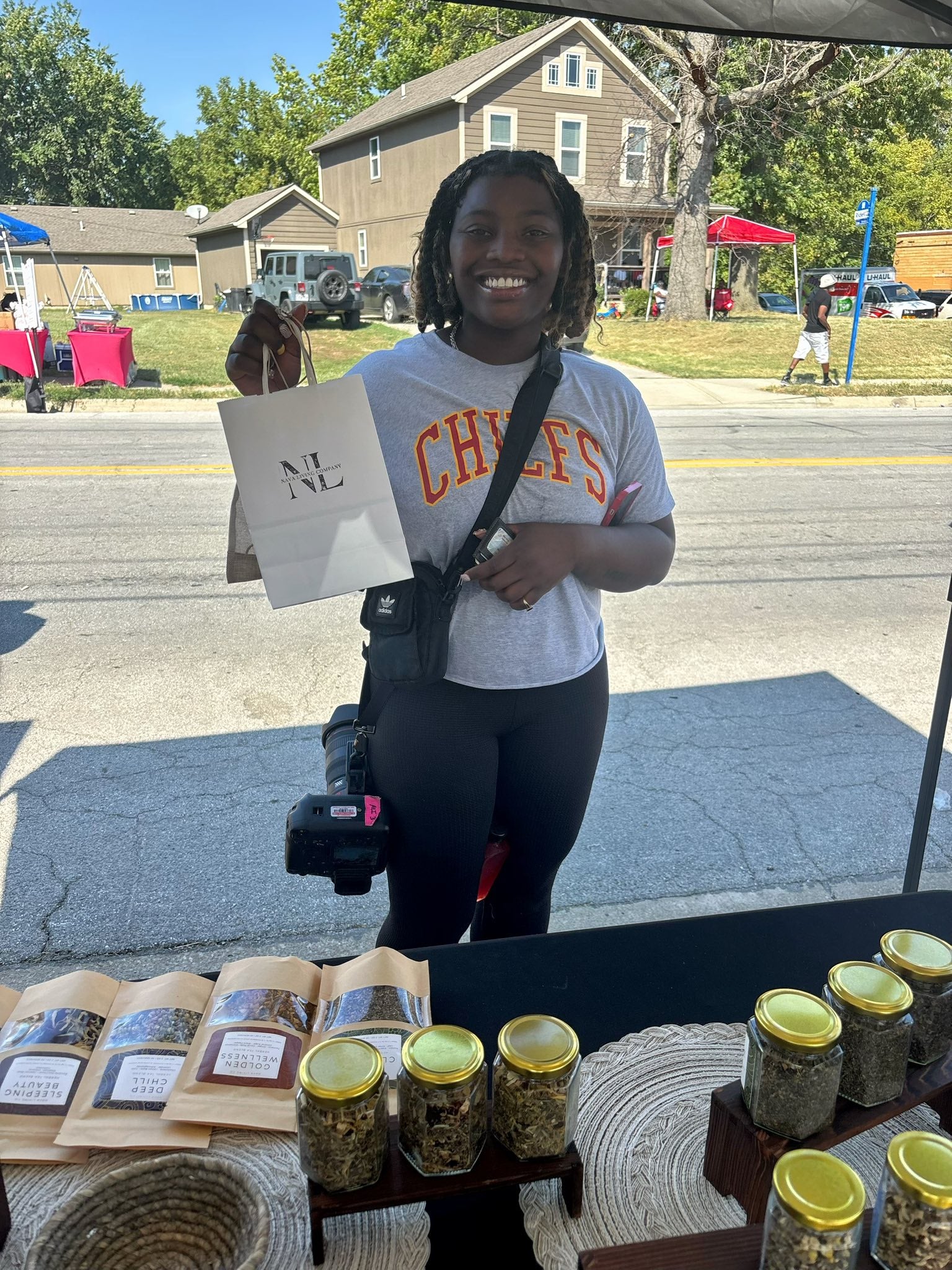 Person holding a Nava Living Co herbal tea blend product at an outdoor market with loose leaf tea jars and packets on a table.