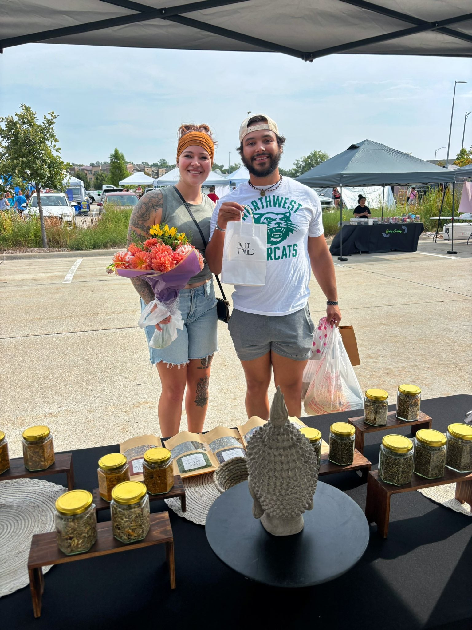 Two people standing behind a table with loose leaf tea jars and a statue, with a parking lot and tents in the background.