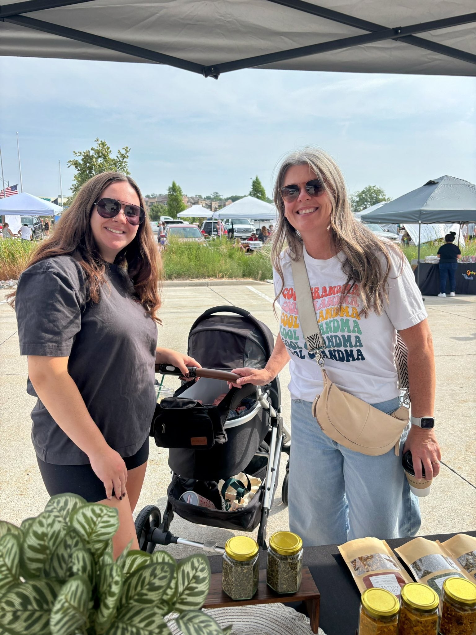 Two women standing next to a stroller with Nava Living Co. loose leaf herbal tea jars on a table outdoors.