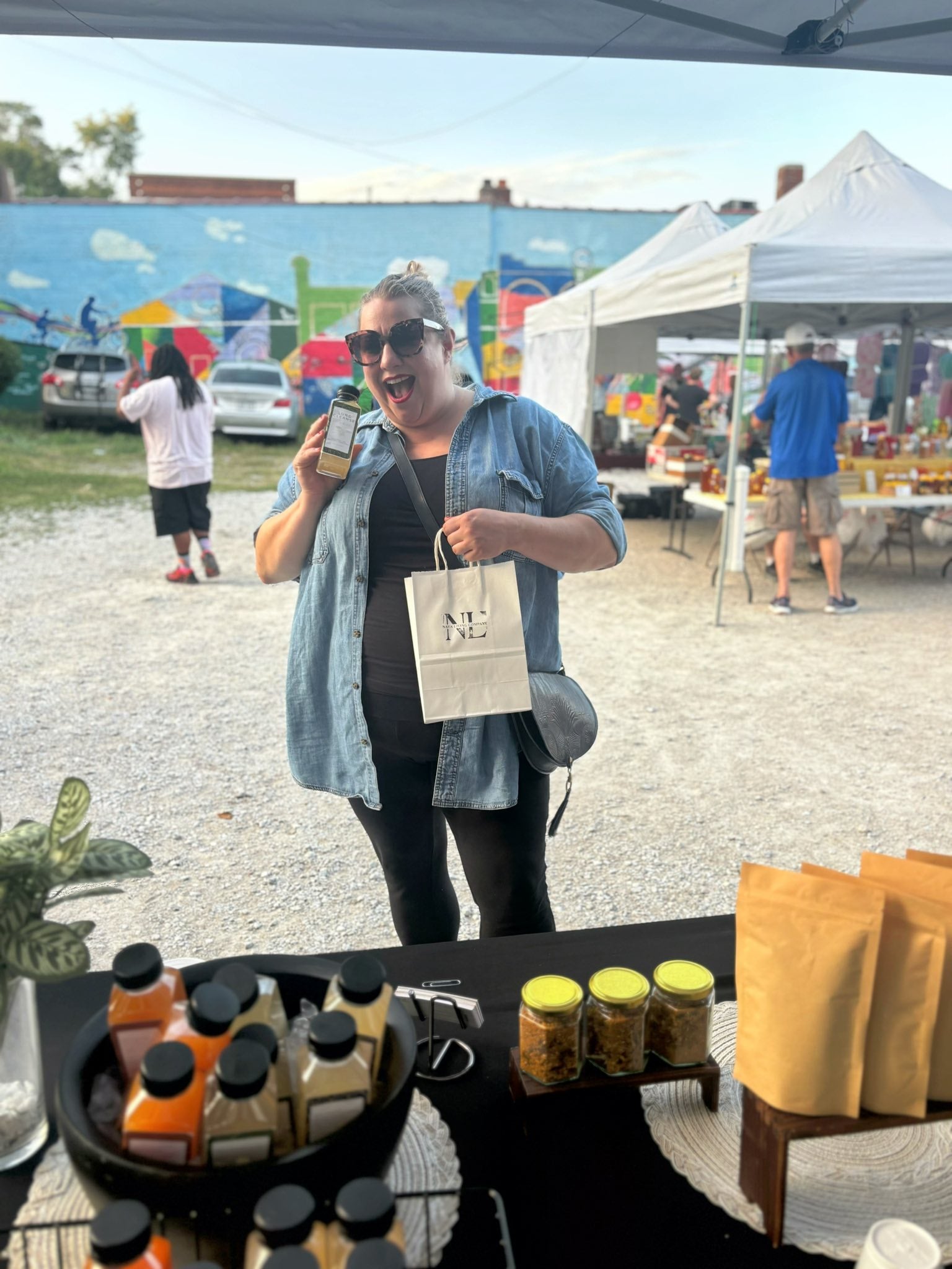 Woman holding a Nava Living Co. loose leaf tea bag at an outdoor event with food and drink items on a table.