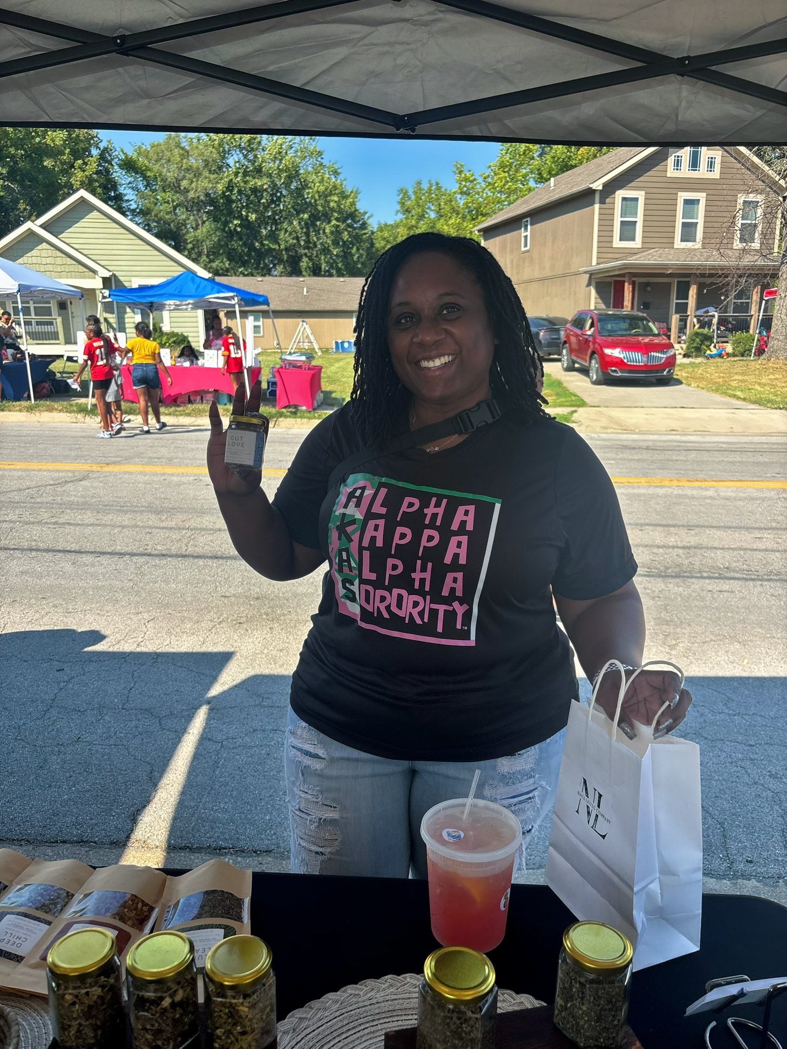 Woman in a 'Alpha Gamma Delta' shirt standing behind a table with Nava Living Co loose leaf tea jars and a drink, outdoors.