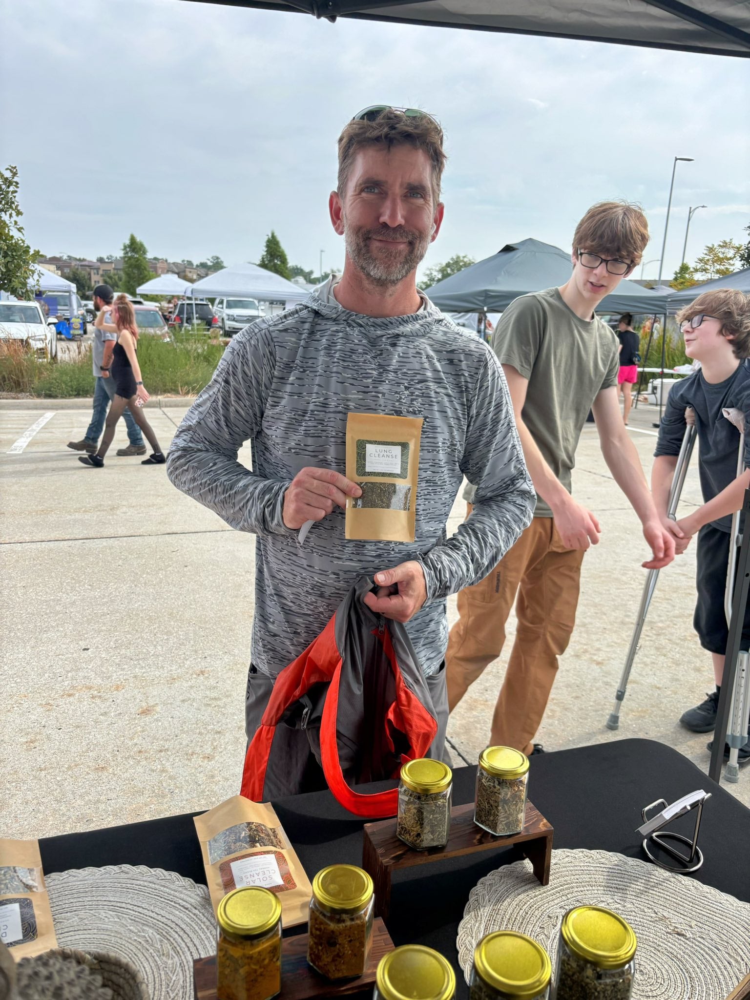 Man holding a Nava Living Co. herbal tea product package at an outdoor market with loose-leaf tea filled jars on a table.